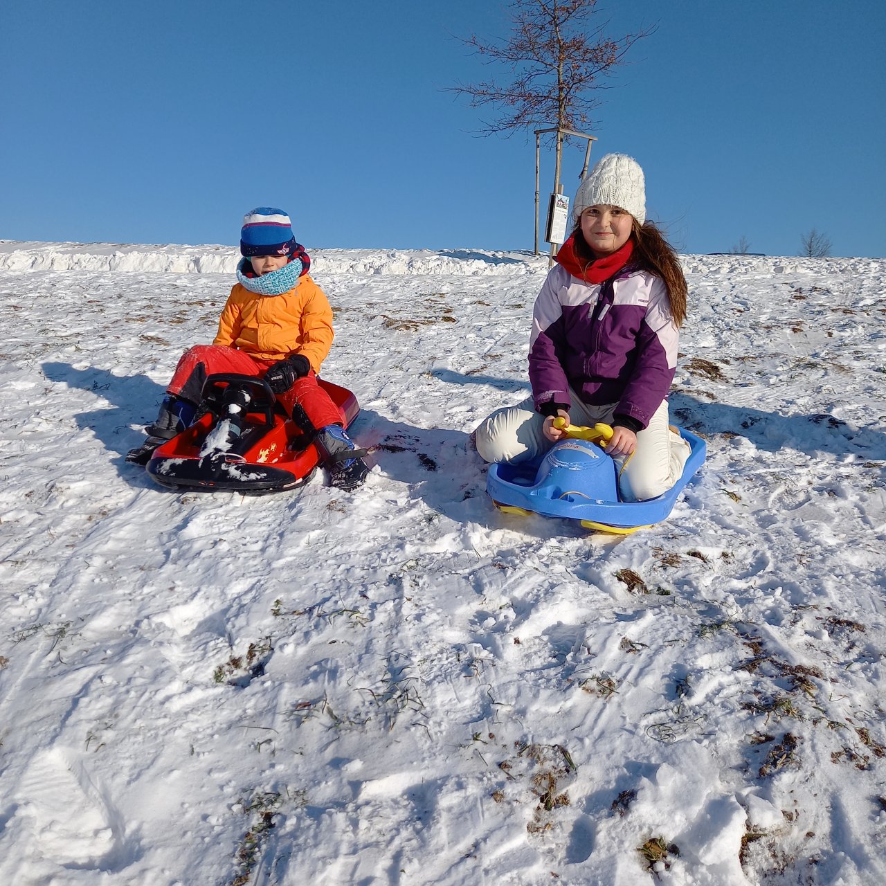 Zwei Kinder sind auf ihren Schlitten und fahren den Berg hinunterbei strahlend blauem Himmel.