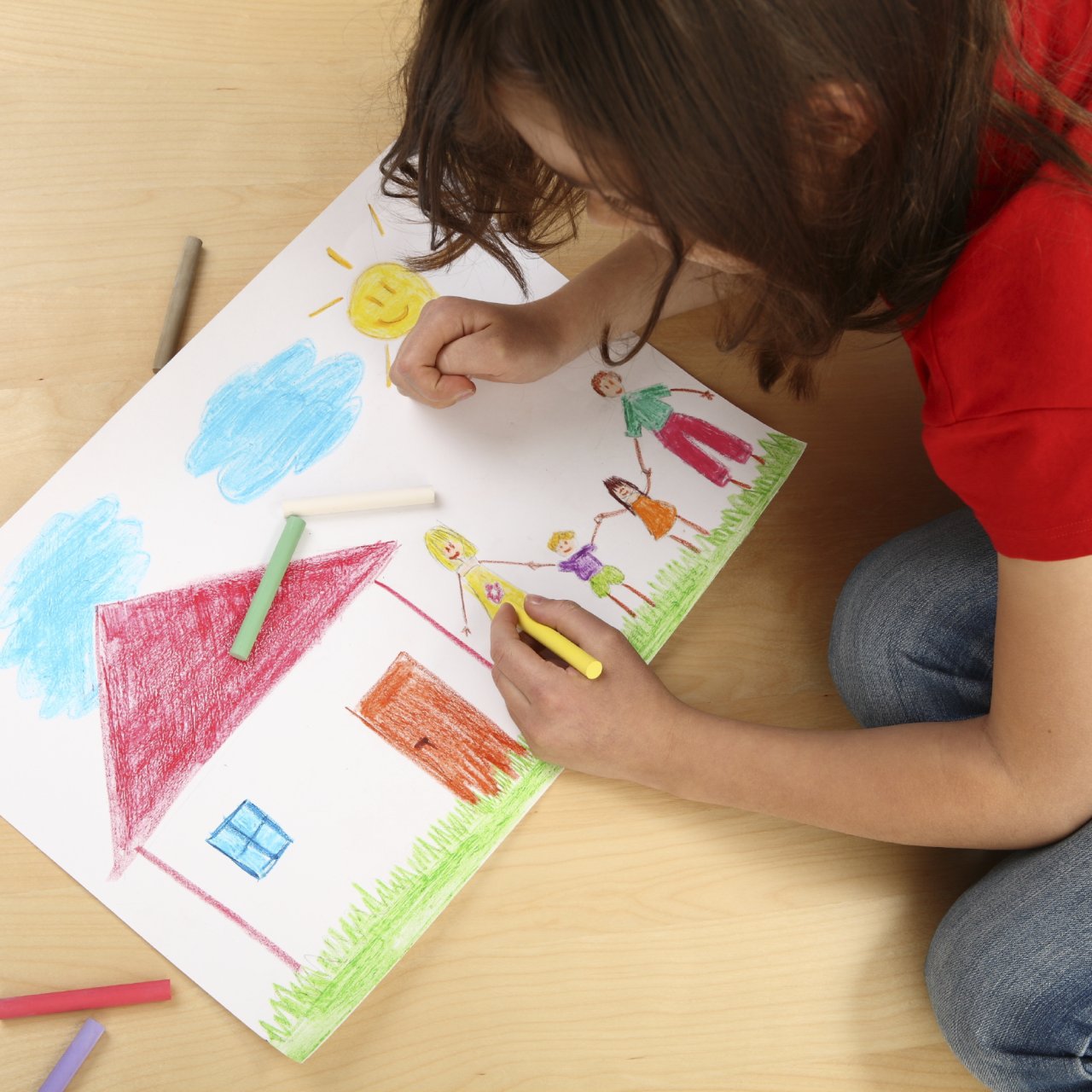 Mädchen malt auf einem weissen Papier ein schönes farbiges Bild mit Wachsmalstiften. Auf dem Bild hat es ein Haus mit einer 4-köpfigen Familie, eine Sonne und zwei Wolken.