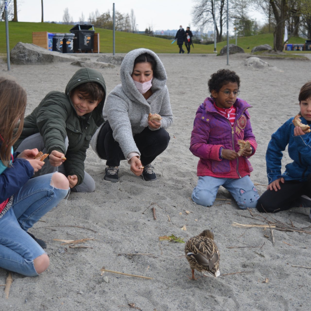 Gruppe mit Kindern und Jugendlichen füttert eine Ente am See. 